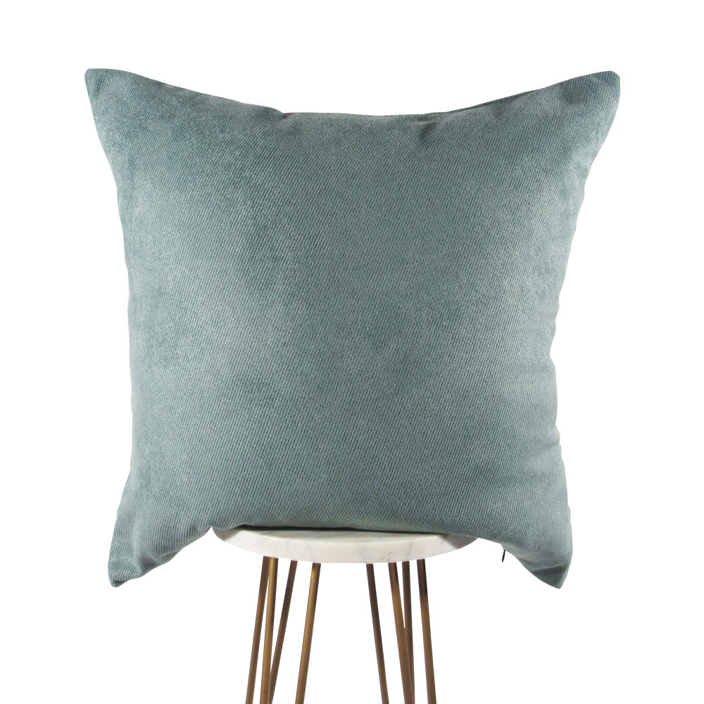 A square-shaped, green throw pillow with a diagonal indented line pattern, placed on a tripod floor stand.