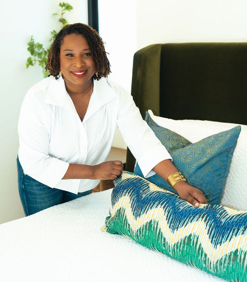 Woman arranging decorative pillows on a bed in a bright room.
