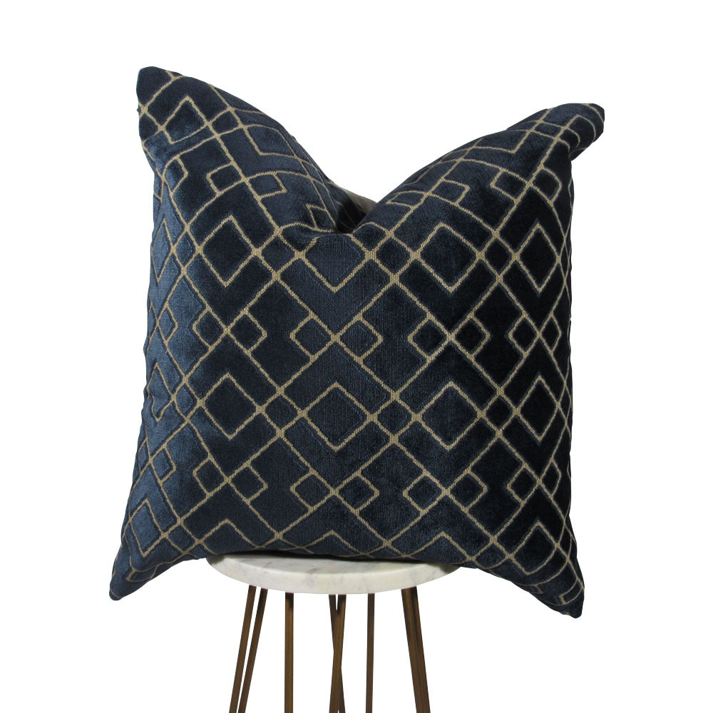 A midnight blue velvet pillow with a geometric beige pattern, displayed on a simple wooden stand.