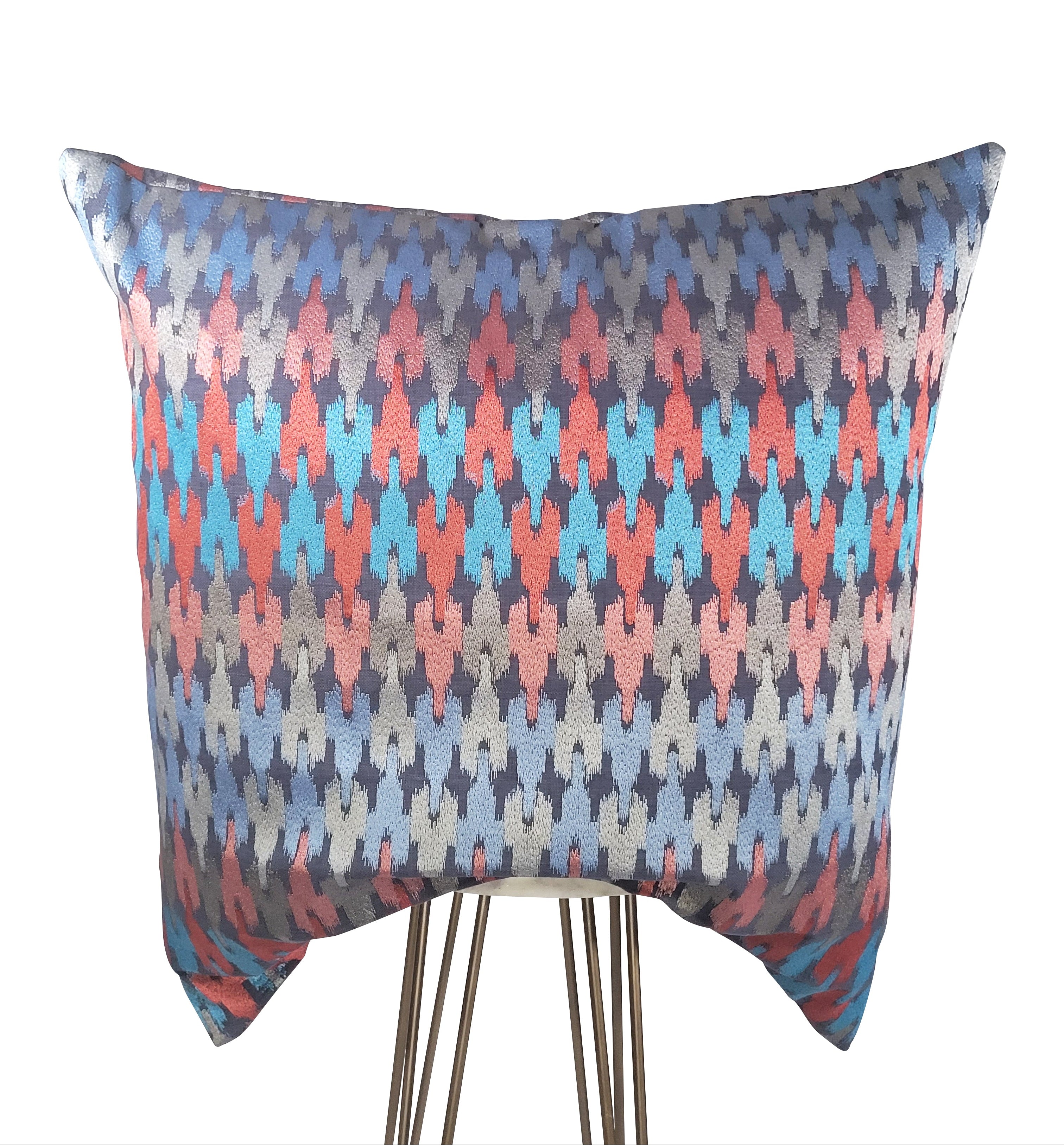 A square-shaped decorative pillow with a geometric pattern featuring blue, red, pink, and silver colors, and a slight sheen to the fabric.